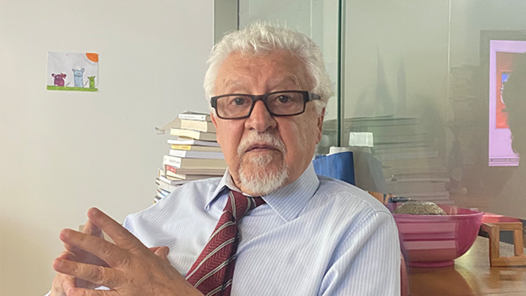 Fernando Mitre de camisa social azul e gravata vermelha está sentado em um escritório, com pilhas de livros ao fundo. Ele usa óculos e olha diretamente para a câmera, com as mãos entrelaçadas à frente.