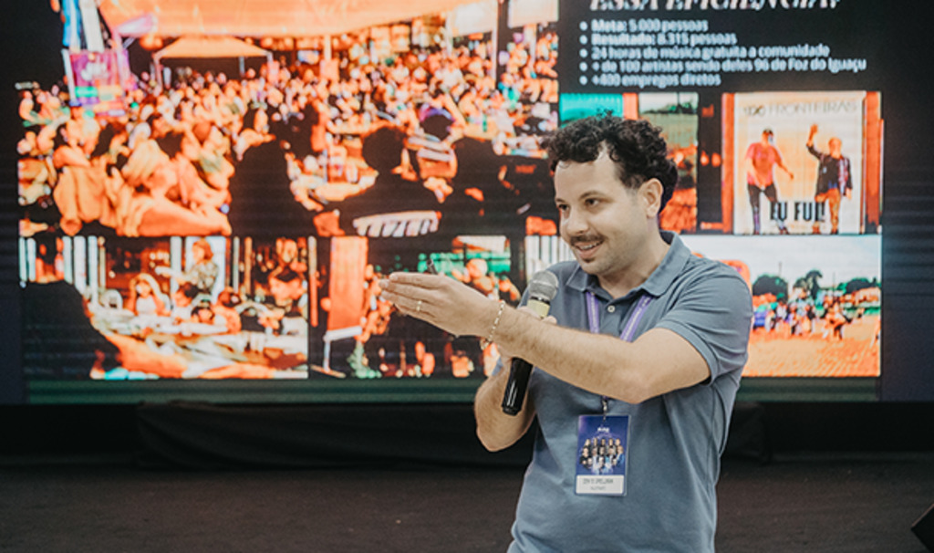 Homem em pé, falando ao microfone, gesticulando com uma das mãos durante uma apresentação. Ele usa camiseta polo azul e crachá. Ao fundo, há um grande telão com imagens de um evento com público e slides, sugerindo uma palestra ou conferência em andamento.