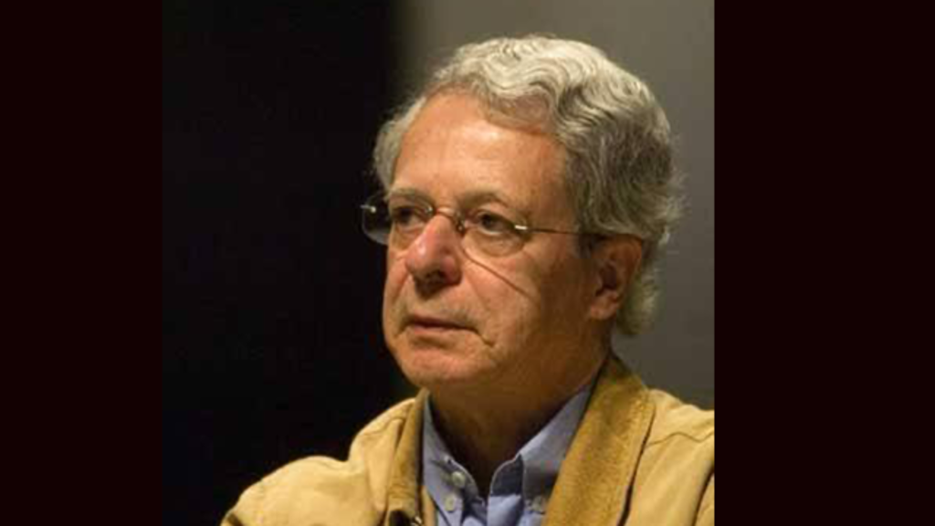 A imagem mostra um homem mais velho, de cabelos grisalhos e curtos, usando óculos. Ele veste uma jaqueta bege sobre uma camisa azul. O homem está sentado e parece estar em um ambiente interno, com expressão séria e olhar voltado para frente.