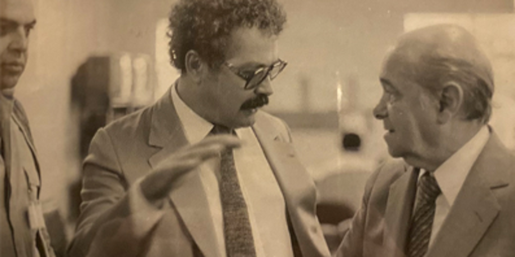 Foto em preto e branco mostra homens de terno conversando. O homem ao centro gesticula com a mão enquanto fala com o homem à direita; ambos parecem envolvidos na conversa.