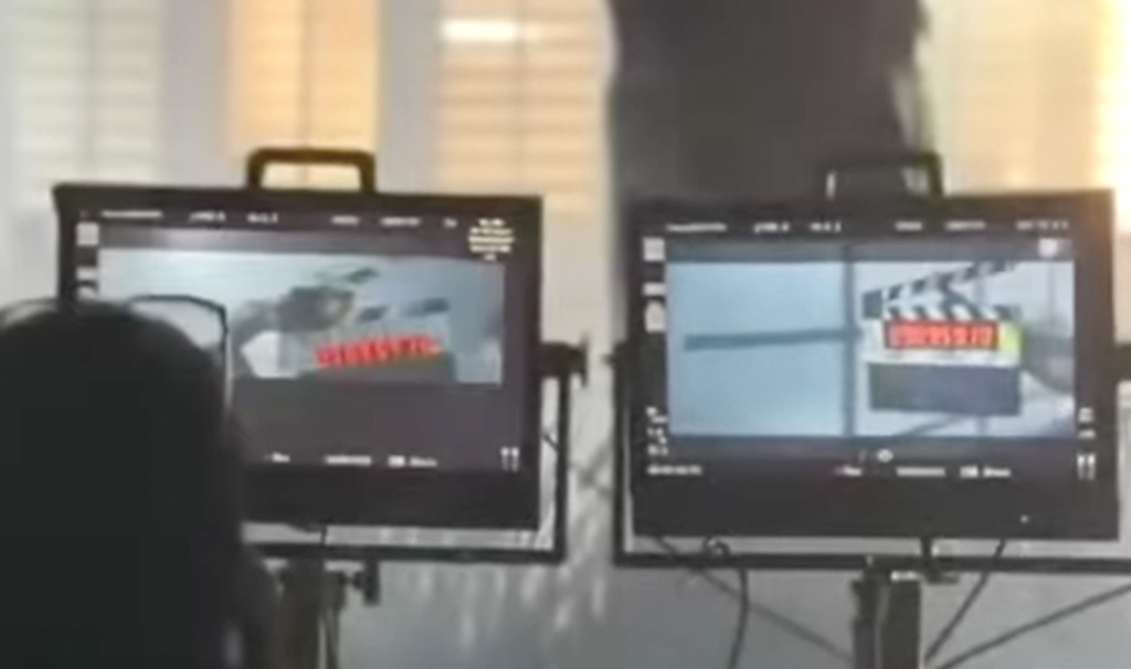 Imagem mostra dois monitores de vídeo profissionais lado a lado, apoiados em suportes metálicos. Em cada monitor aparece a imagem de uma claquete de cinema, indicando uma gravação audiovisual em andamento. Os monitores estão em um ambiente interno, com luz entrando por uma janela ao fundo. À esquerda, vê-se parcialmente a silhueta desfocada de uma pessoa, de costas para a câmera, observando os monitores. Cabos estão conectados aos equipamentos, reforçando o contexto de um set de filmagem ou estúdio de produção.