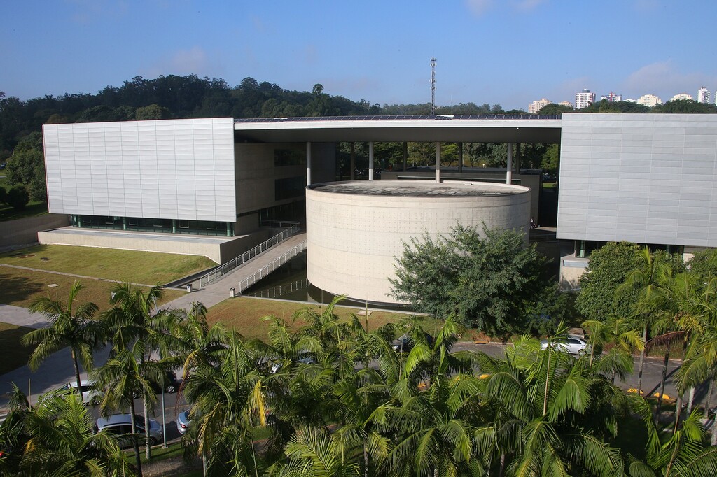 Fotografia aérea da Biblioteca Brasiliana Guita e José Mindlin (BBM), localizada na Universidade de São Paulo (USP). A imagem mostra o edifício moderno com estrutura de linhas retas e fachada metálica cinza, interligando dois blocos retangulares por uma passarela sobre um espelho d’água. No centro, há uma construção cilíndrica de concreto, parcialmente sombreada por uma ampla cobertura plana sustentada por pilares. Em primeiro plano, há fileiras de palmeiras e carros estacionados, enquanto ao fundo vê-se uma área arborizada e prédios residenciais ao longe, sob um céu azul claro.